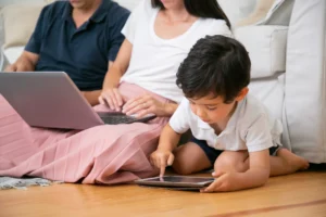 A focused little boy using tablet by himself sitting floor living room by his parents with laptop.